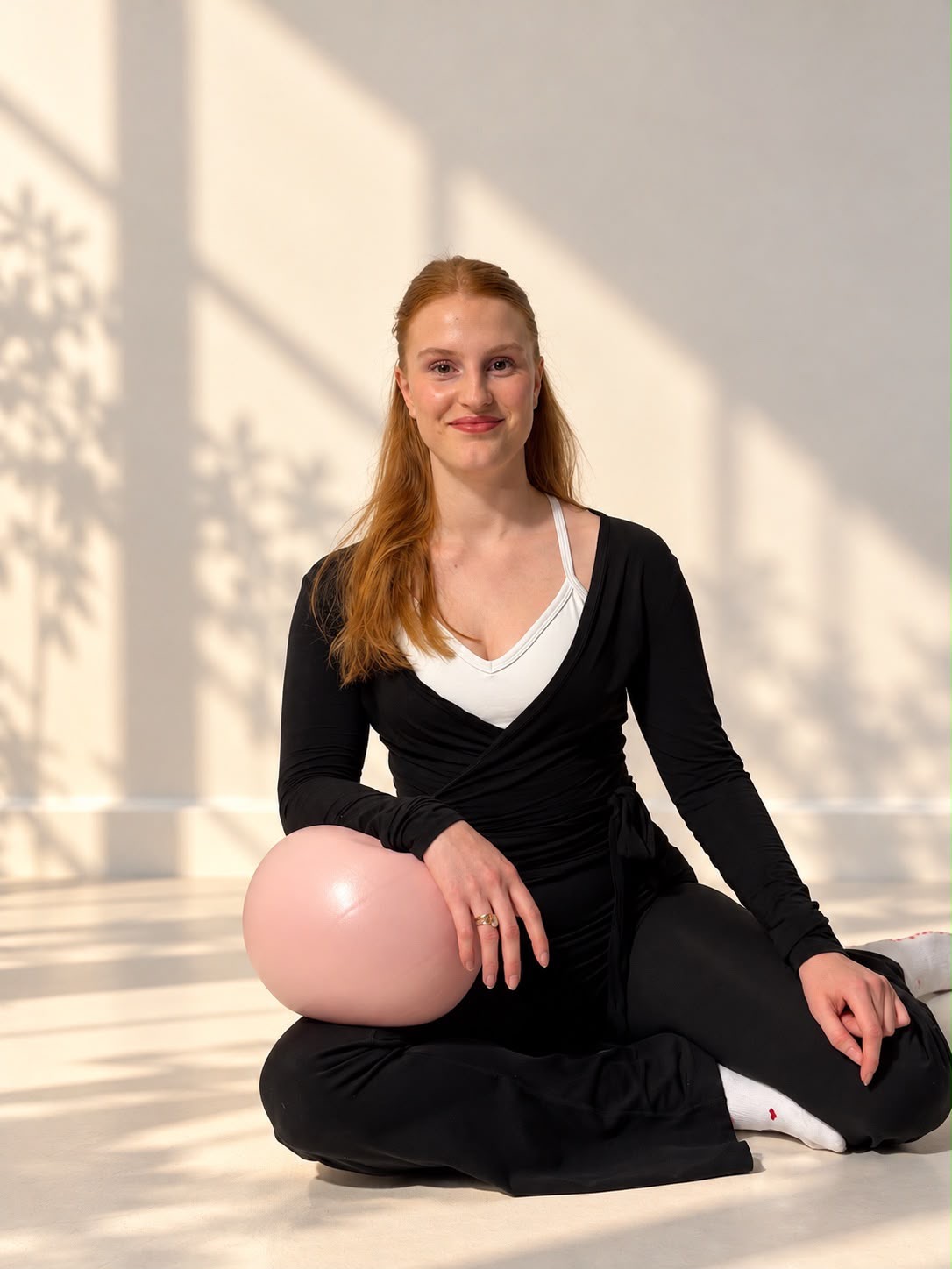 Selina sitzt lächelnd mit rosa Pilates-Ball auf der Matte